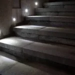 A series of step lights inlaid into a wall next to a concrete staircase, one light illuminating each step.